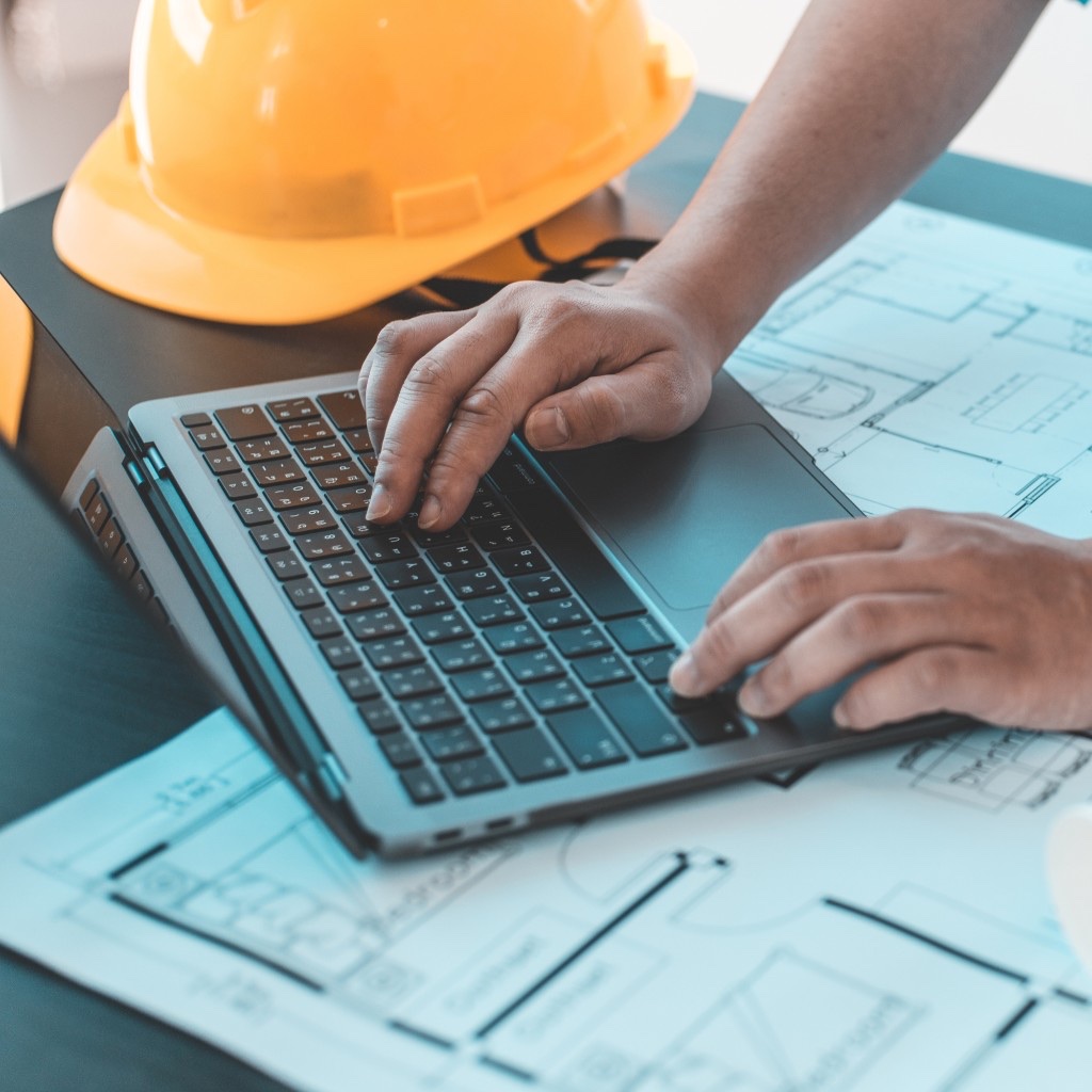 An engineer working on a laptop with blueprints and a hard hat on the desk